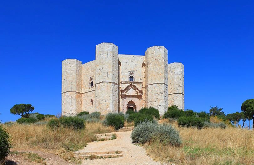 Castel del Monte visto frontalmente, circondato dalla vegetazione sotto un cielo blu intenso.