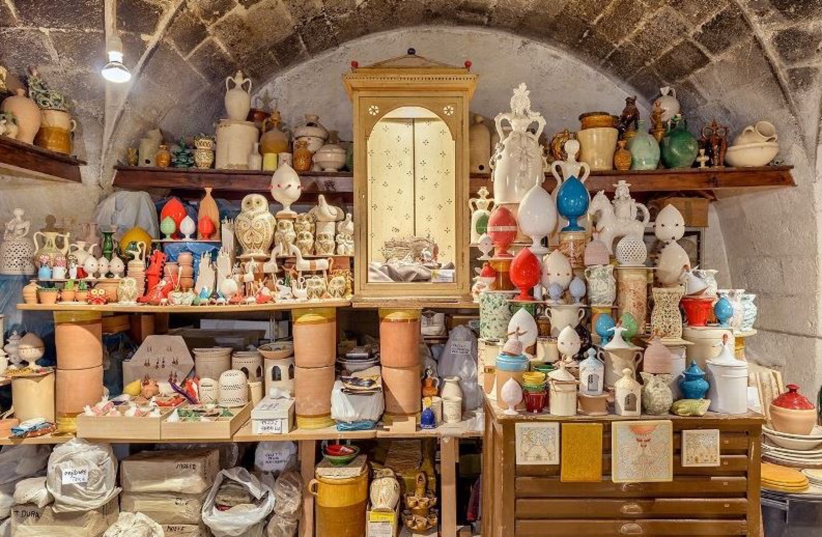 Una bottega voltata in pietra colma di ceramiche di Grottaglie: pumi, vasi e figure sui ripiani di legno.